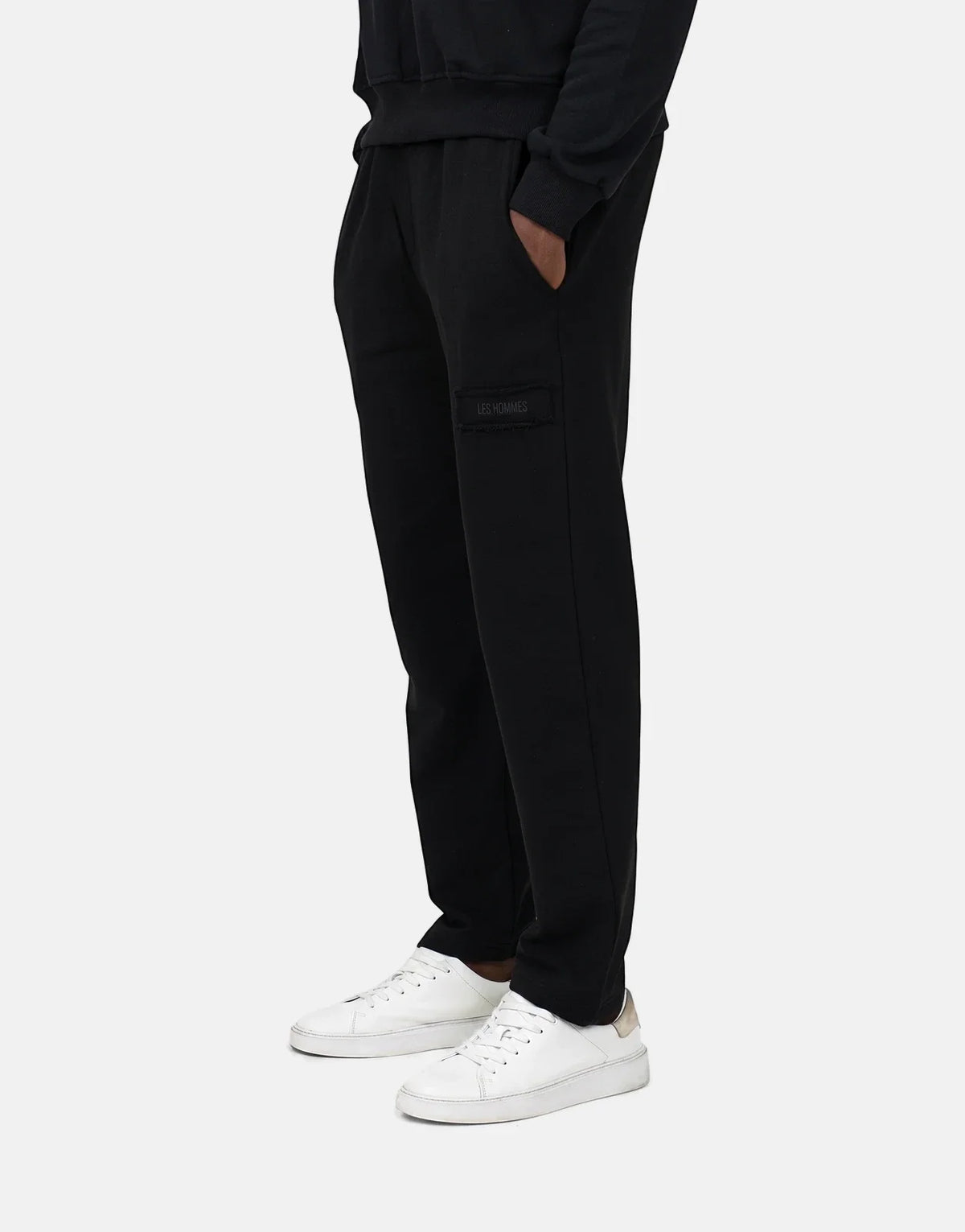 Men's Les Hommes black fleece joggers, seen from a side view, featuring a logo patch and worn with white sneakers.