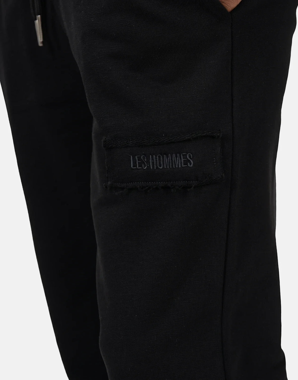 Close-up of Les Hommes black jogger pants featuring a stitched logo label and a silver drawstring tip, side view.