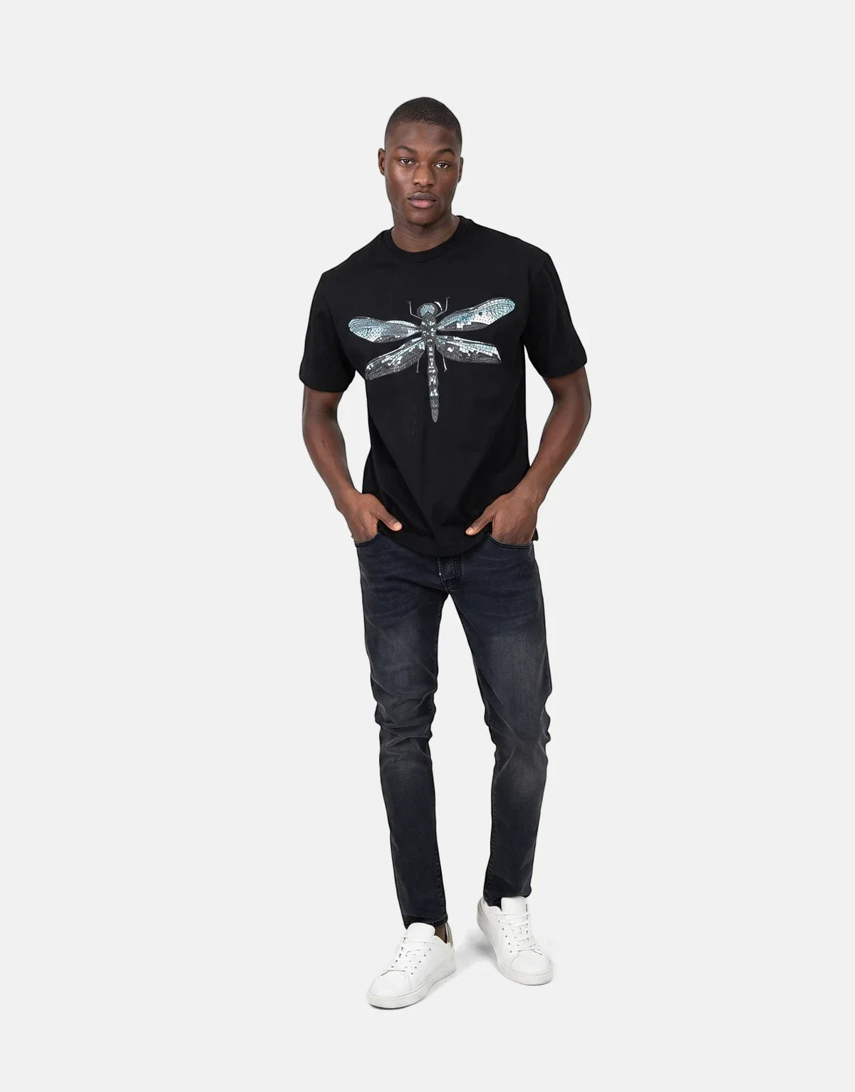 A male model wears a black Les Hommes Dragonfly T-shirt, dark wash jeans, and white sneakers, standing full-body.