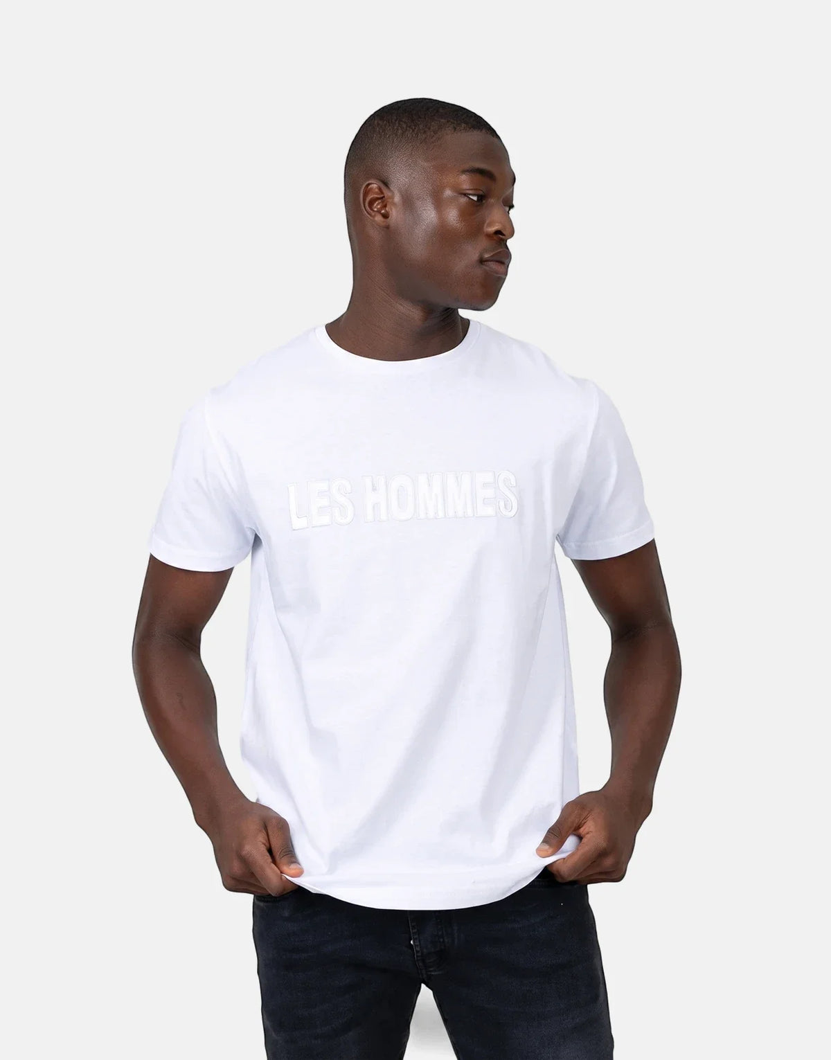 A male model wears a Les Hommes white short-sleeve t-shirt with a glitter logo and dark jeans, looking to the side.