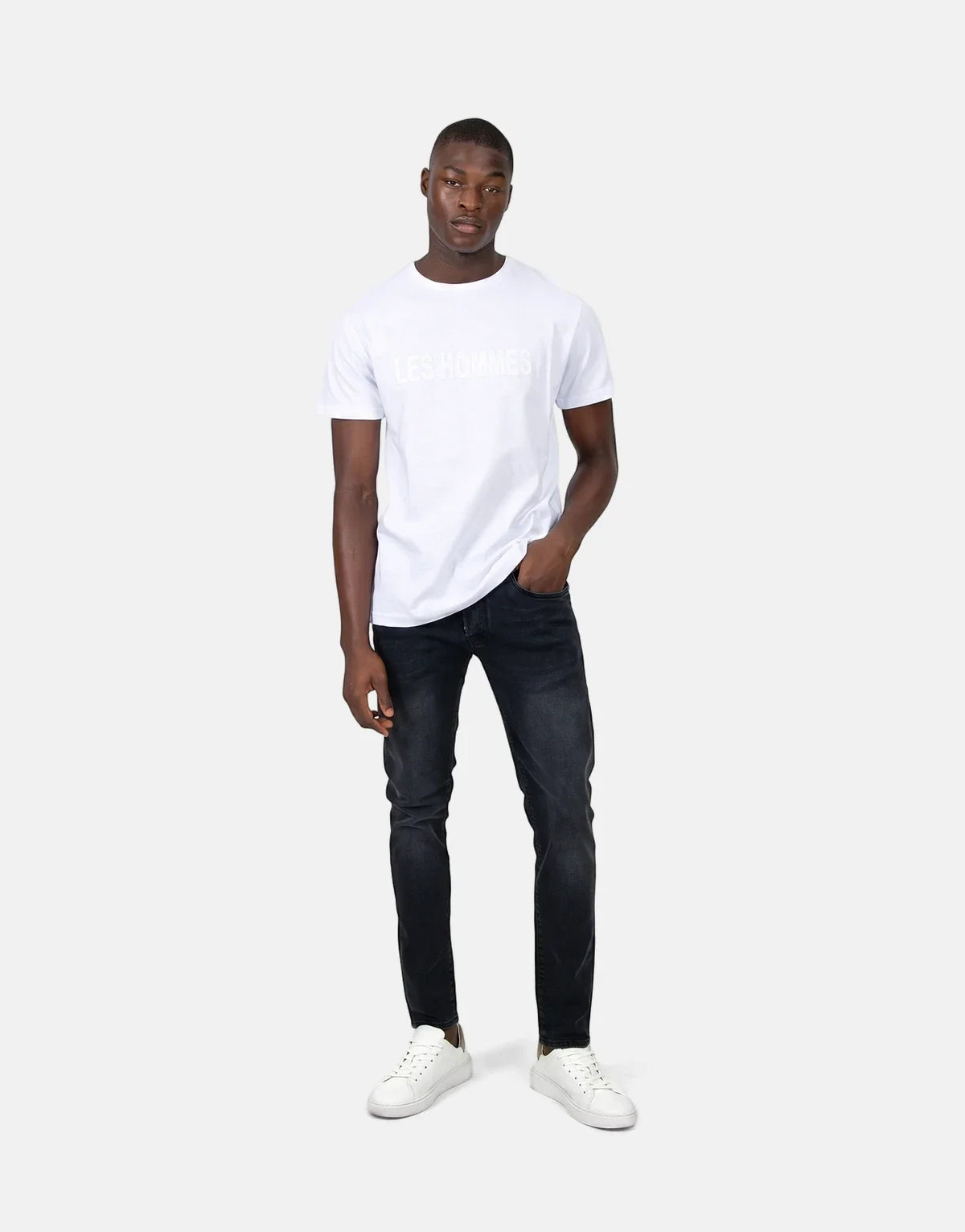 A young man wears a white Les Hommes t-shirt with a glitter logo, black jeans, and white sneakers, posing full-body.
