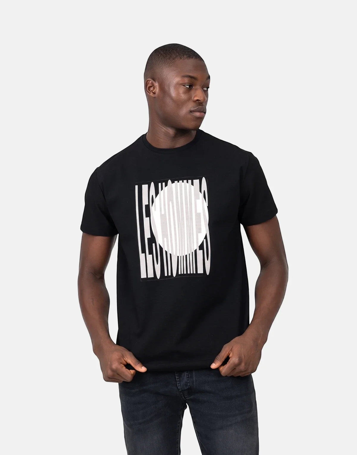 A male model wears a black Les Hommes t-shirt featuring a distinctive white logo print, paired with dark jeans, posed in a