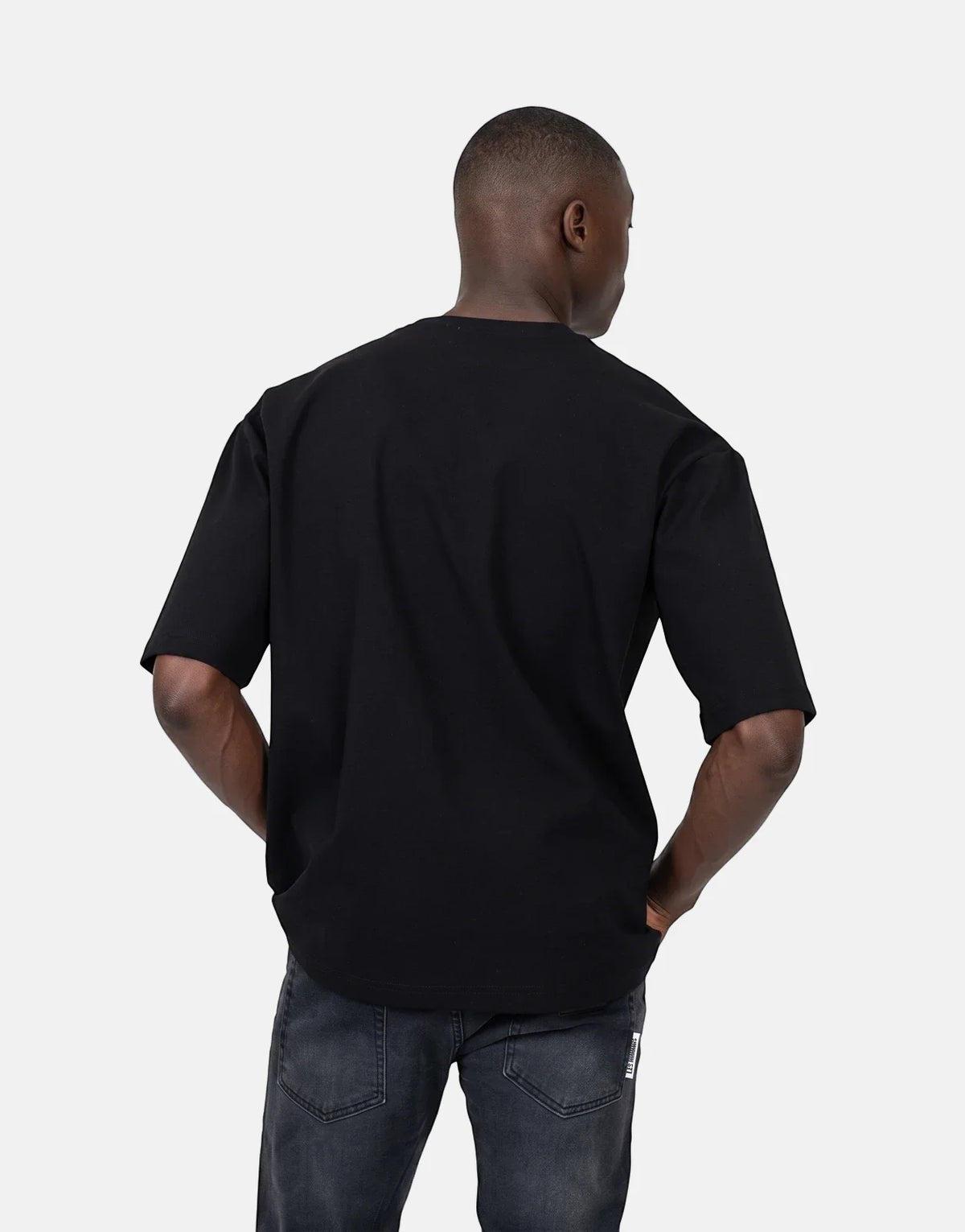 Les Hommes Over Rubber Print Black T-Shirt on a male model, shown from the back with hands in pockets, paired with dark