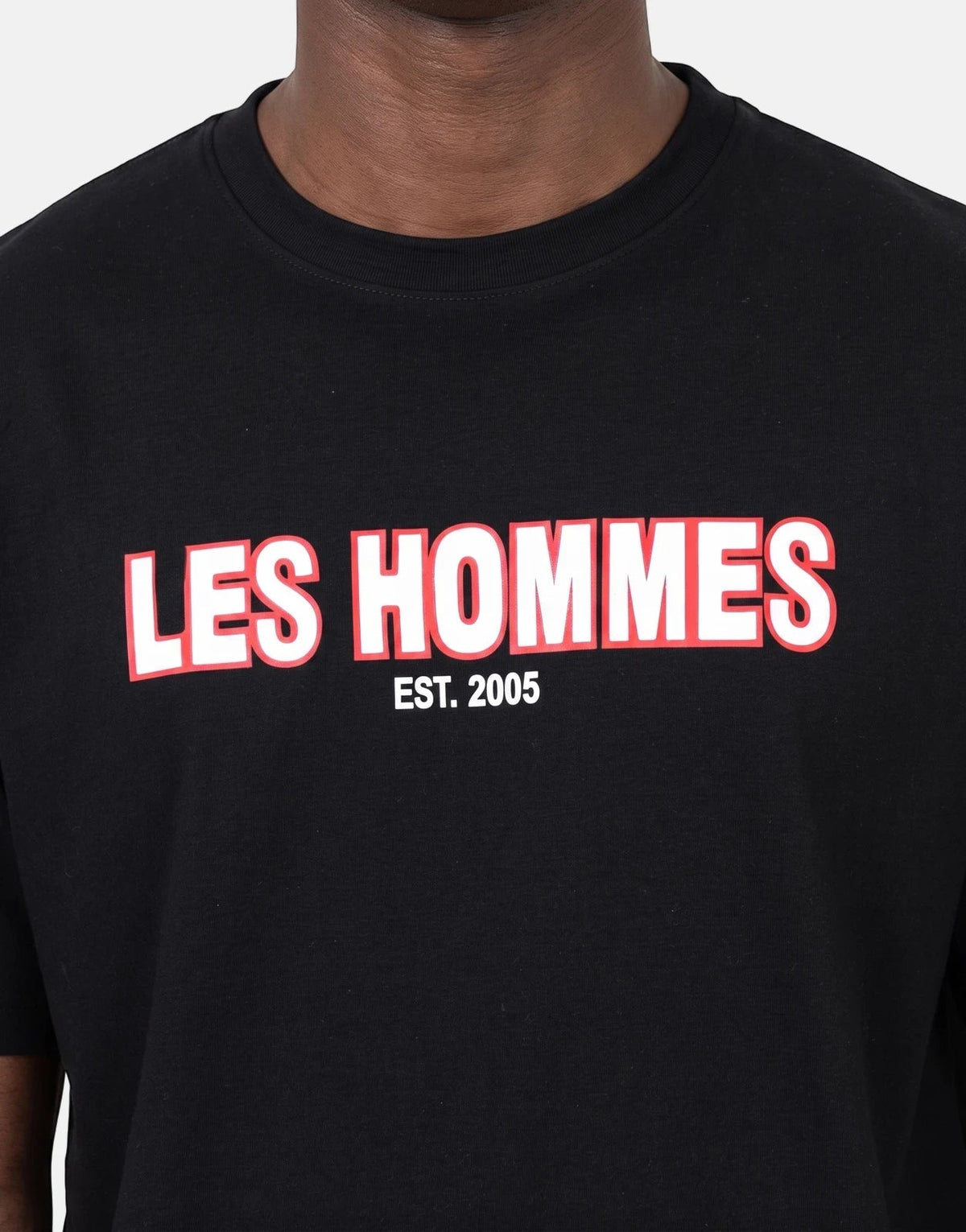 Close-up of a black Les Hommes t-shirt featuring a bold white and red 'LES HOMMES EST. 2005' rubber print, worn by a model.