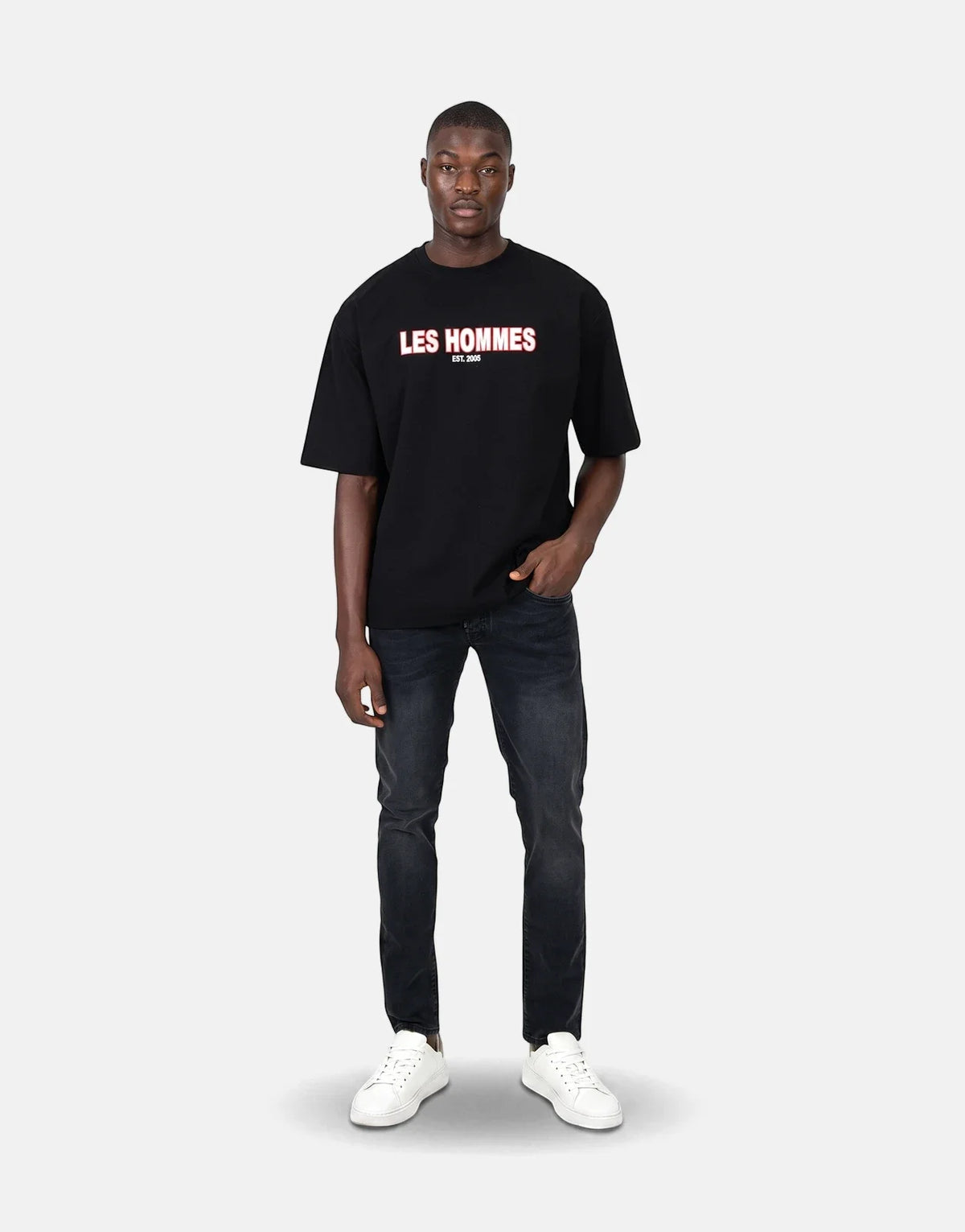 Male model wearing a black Les Hommes Over Rubber Print T-Shirt with black distressed jeans and white sneakers.