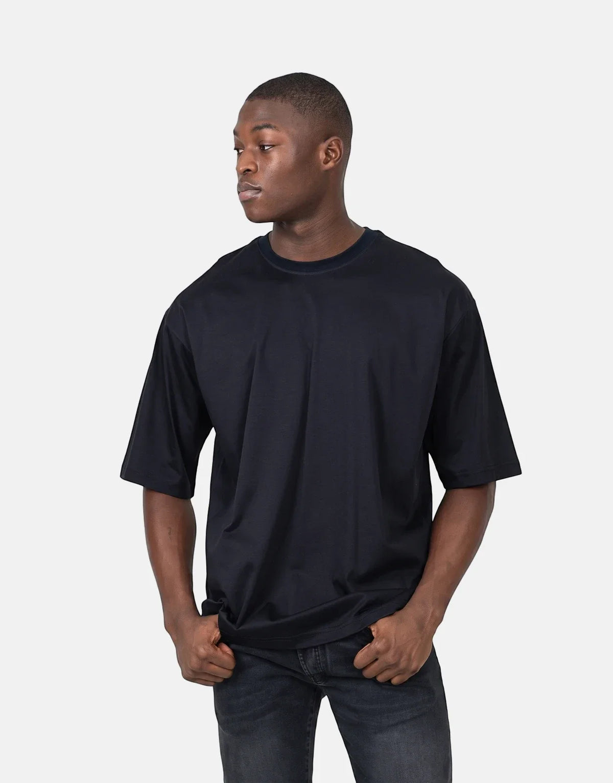 Young man wearing a black Les Hommes oversized t-shirt and dark jeans, hands in pockets, posing against a white background.