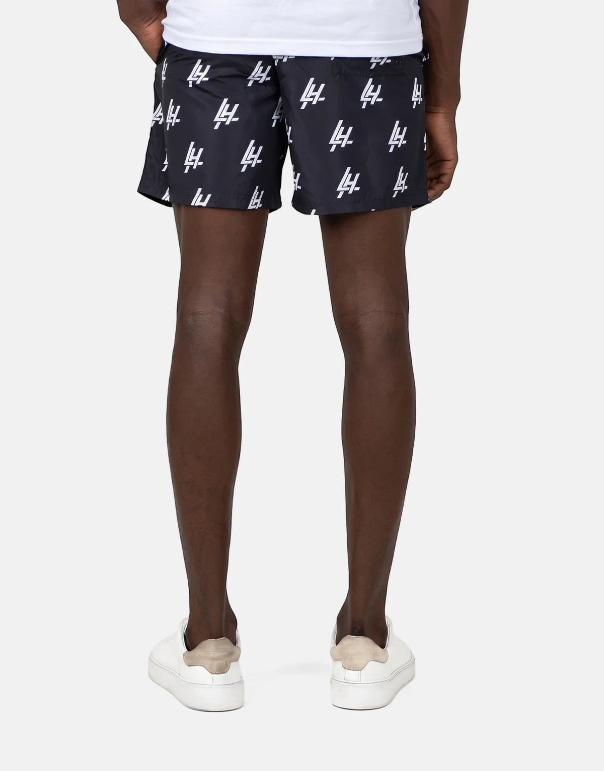 A person's lower body from behind, wearing black Les Hommes swim shorts with white logo print and white sneakers.