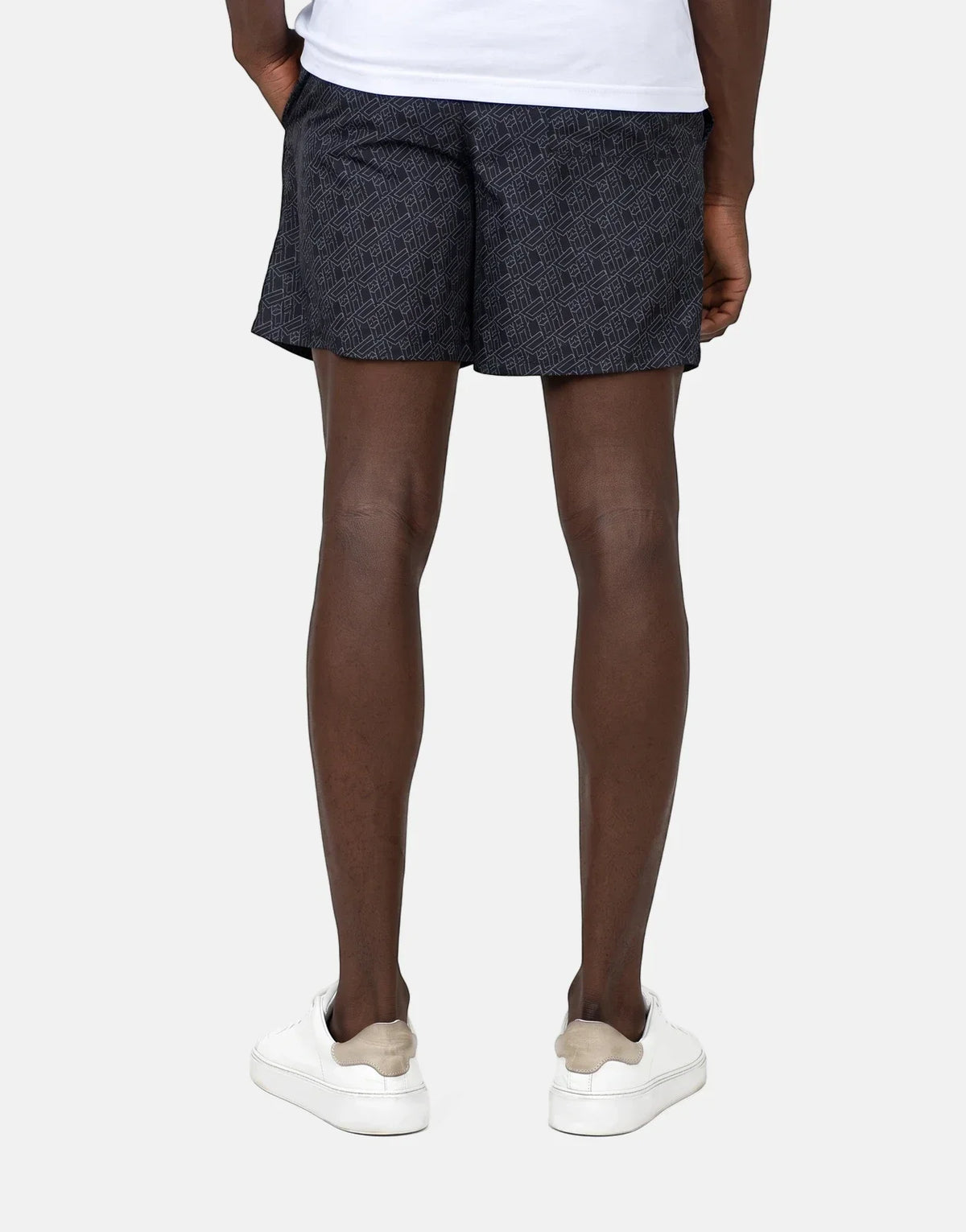 Back view of a person wearing Less Hommes AOP black patterned swim shorts with white sneakers on a simple background.