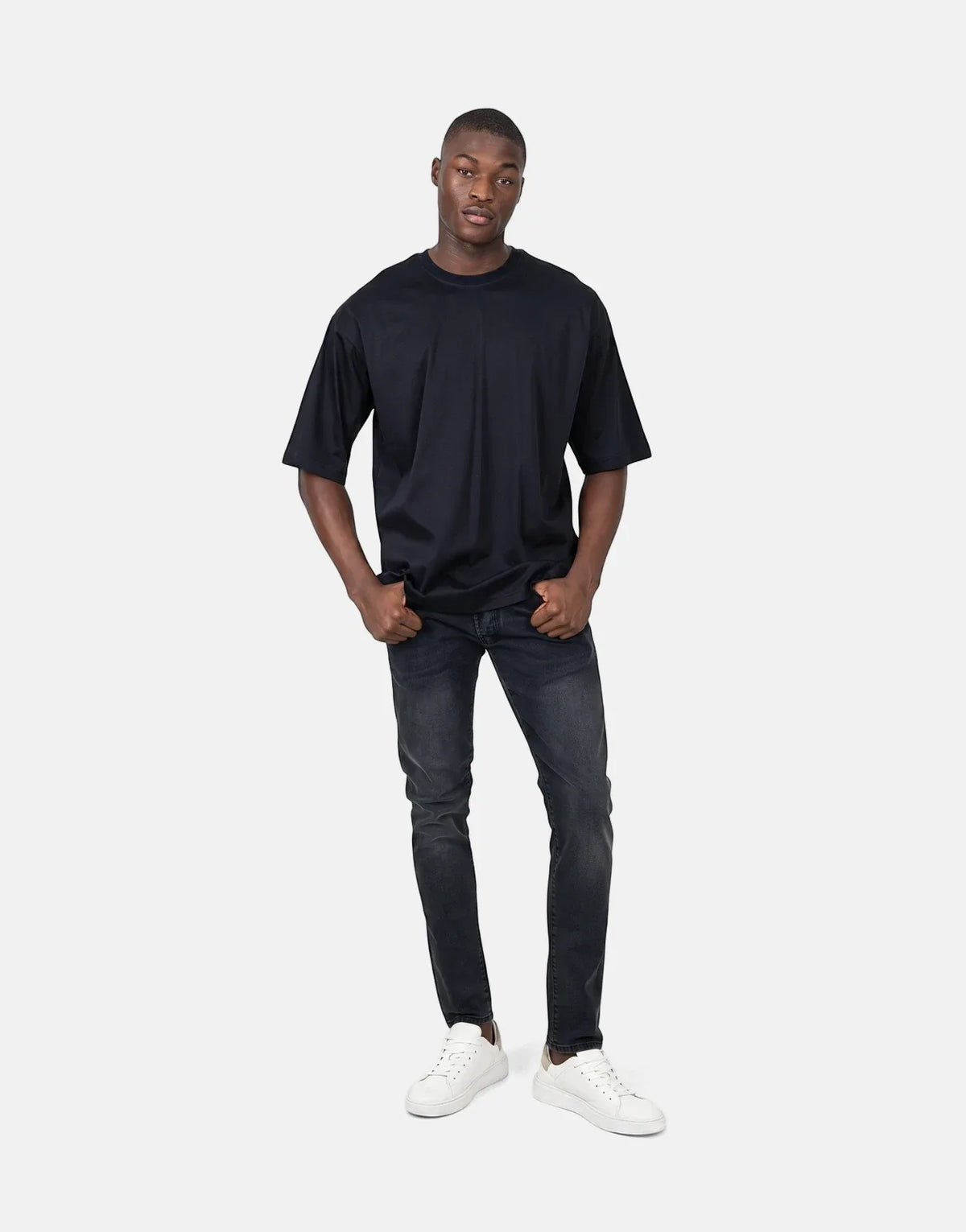 Male model wearing a black oversized t-shirt, black distressed jeans, and white sneakers, standing full body against a white